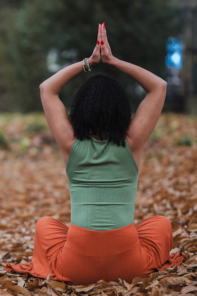 Coach de yoga en lotus, de dos, mains levées vers le ciel, en extérieur.