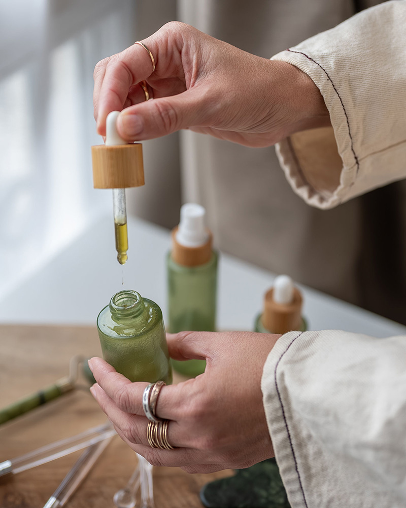 Deux mains tenant un flacon vert et une pipette d'huile faciale dans une ambiance zen et relaxante.