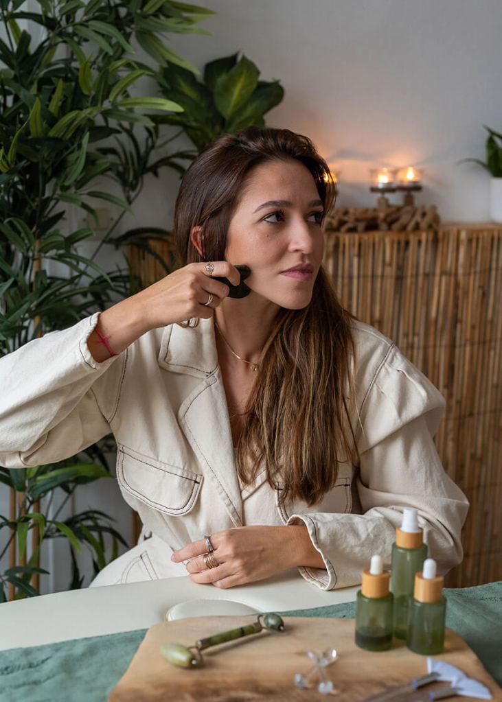 Plan taille d'une jeune femme se massant le visage avec un gua sha dans une ambiance relaxante.