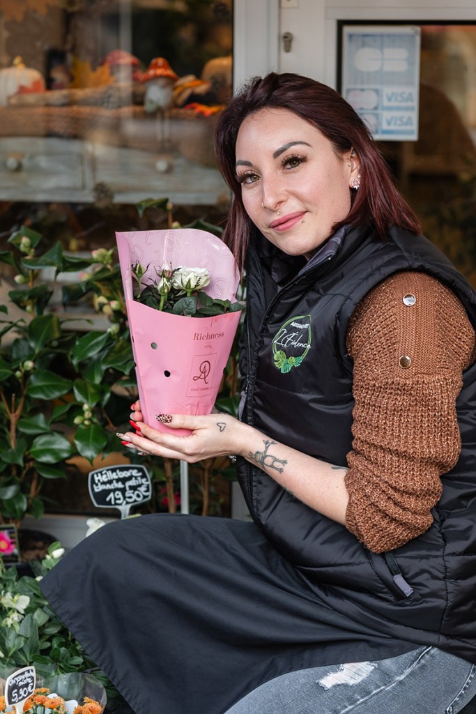 Portrait souriant d'une fleuriste agenouillée devant sa boutique, tenant un petit pot de fleurs.