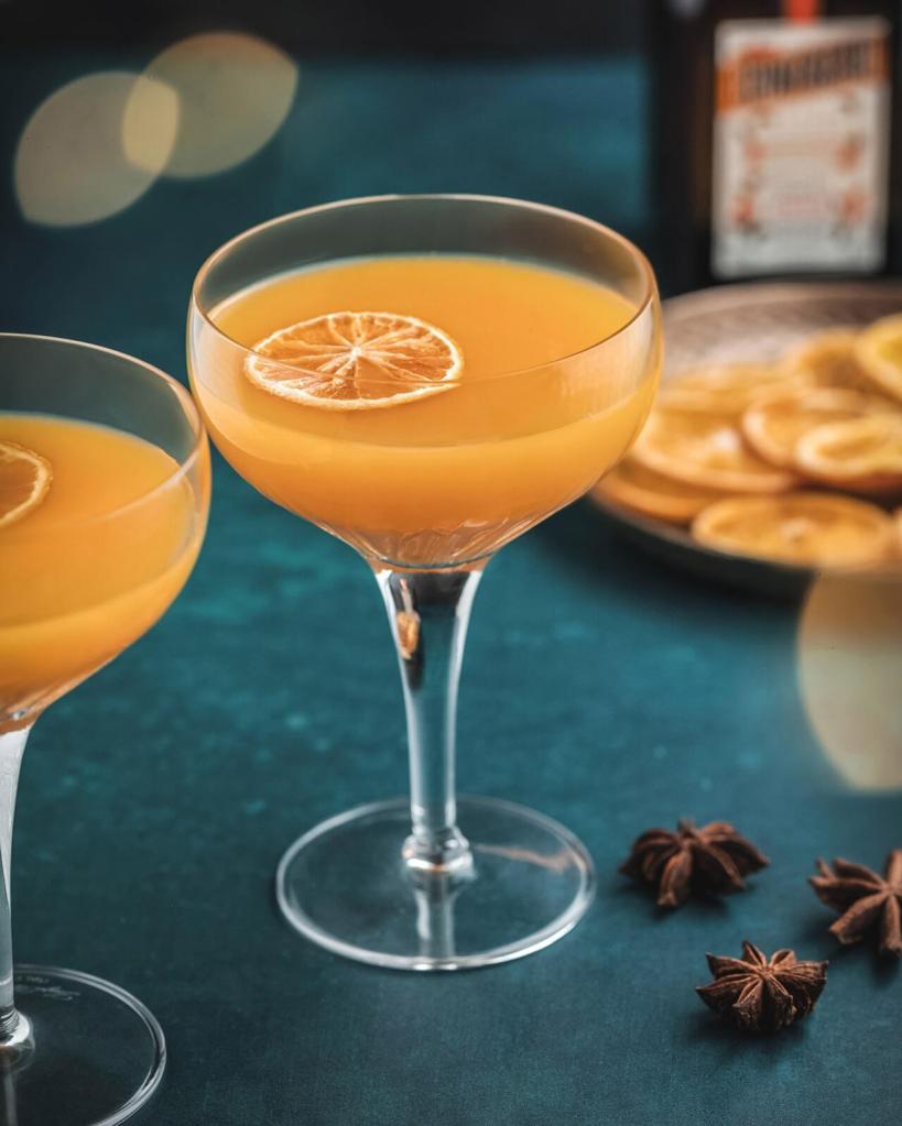 Cocktail orange dans un verre, posé sur une table parsemée d'anis.